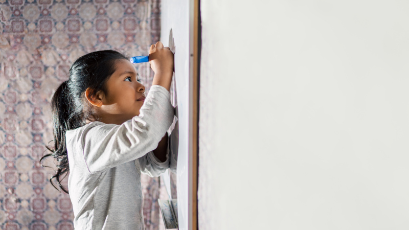 Foto de niña latina sonriente y feliz, escribiendo en la pizarra de clases con plumones de colores.